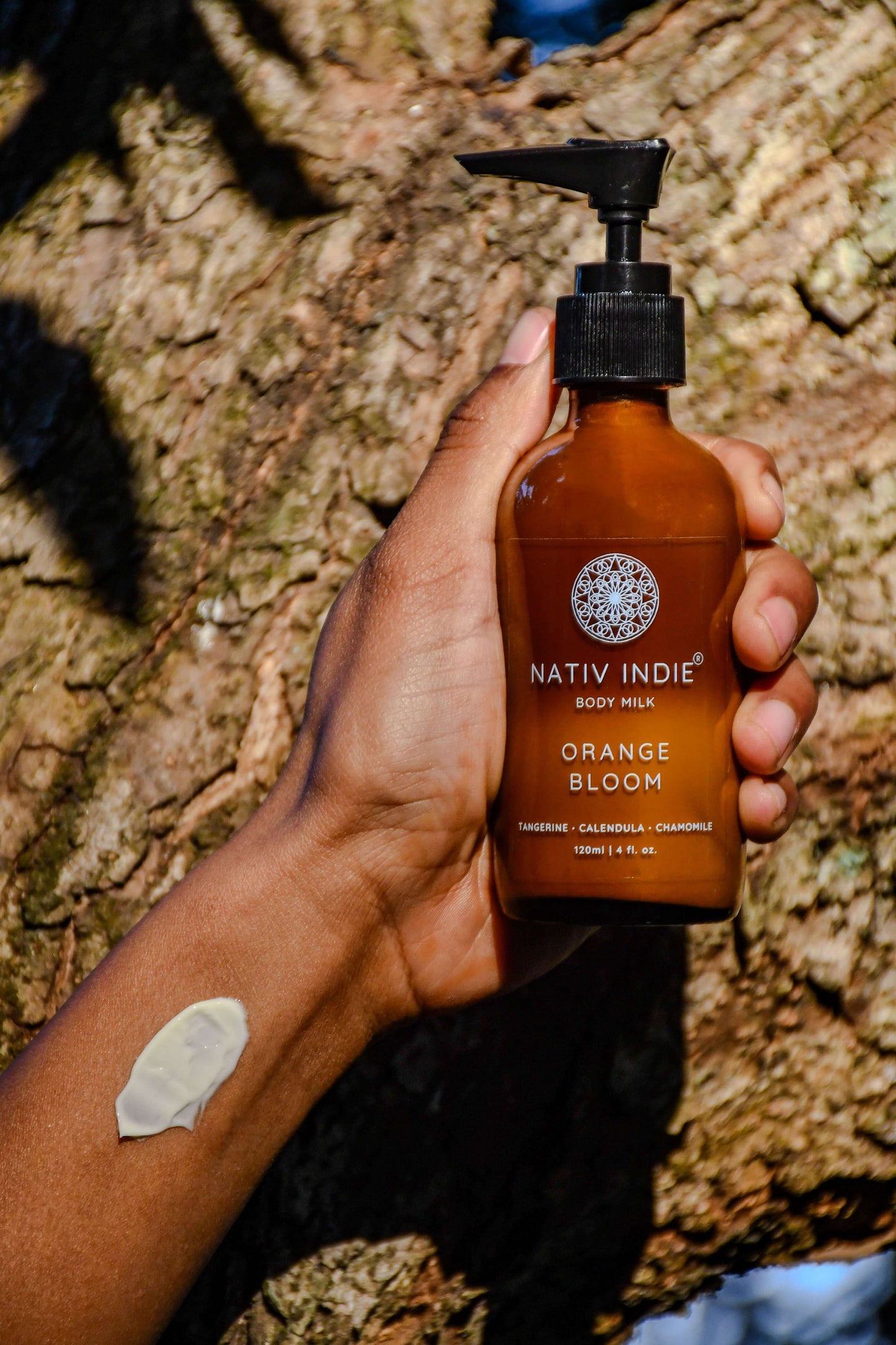 Person holding a bottle of Nativ Indie Orange Bloom Body Milk with a dab of lotion on their arm, nestled against a rustic tree background.